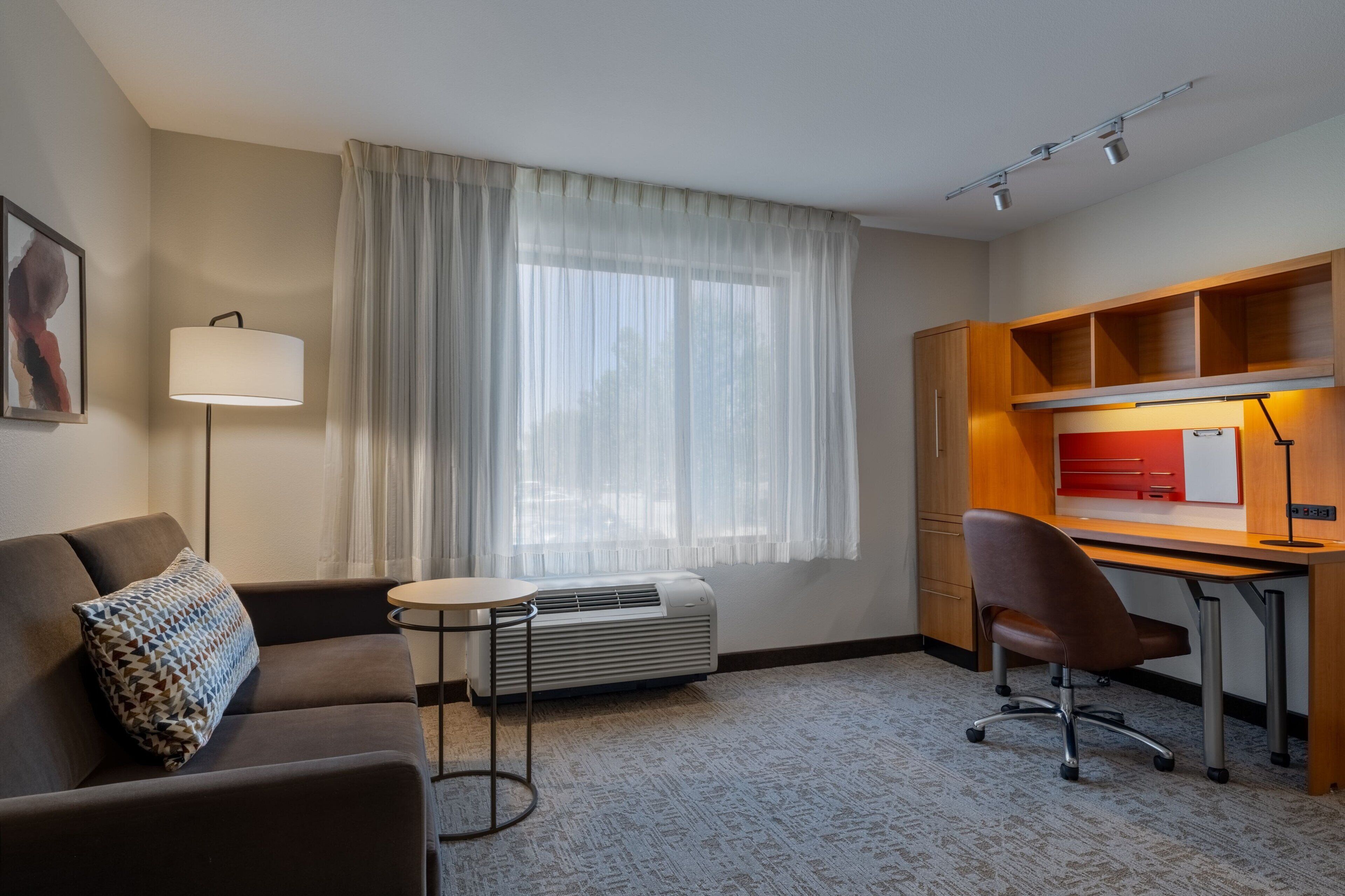 Habitación de hotel con sofá, una pequeña mesa redonda, escritorio con silla, lámpara de pie y ventana con cortinas translúcidas.