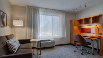 Habitación de hotel con sofá, una pequeña mesa redonda, escritorio con silla, lámpara de pie y ventana con cortinas translúcidas.