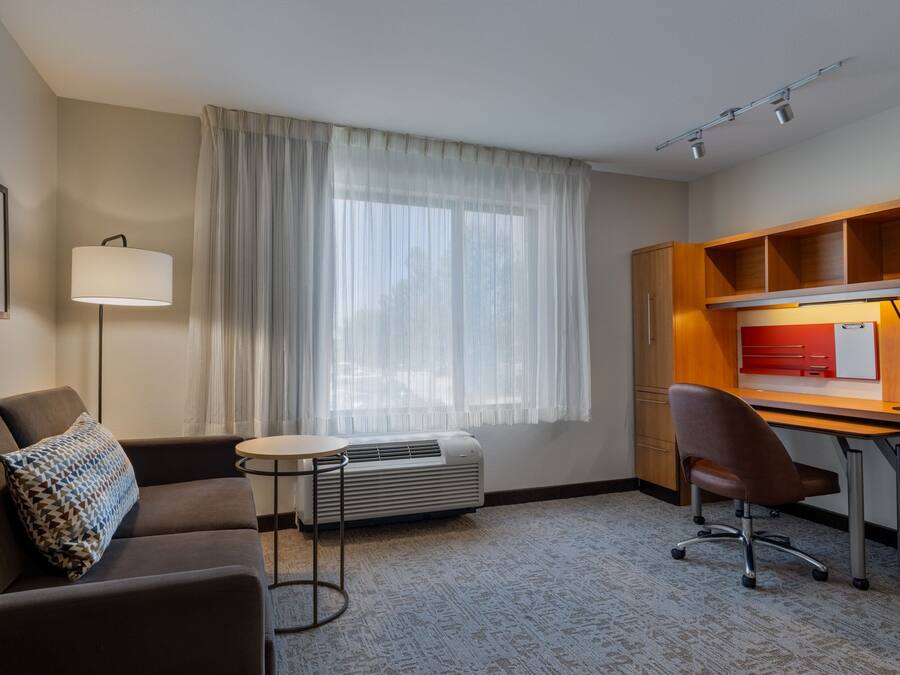 Habitación de hotel con sofá, una pequeña mesa redonda, escritorio con silla, lámpara de pie y ventana con cortinas translúcidas.