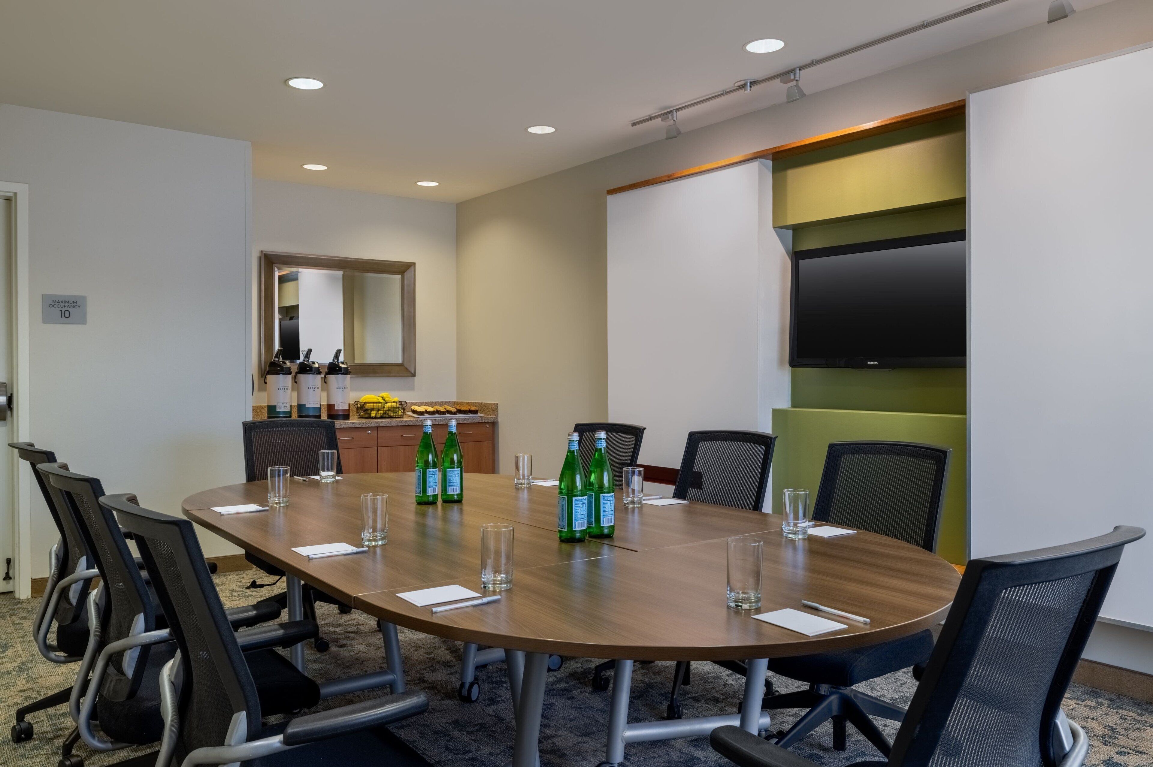 A conference room with a long table, chairs, a flat-screen TV, and a small kitchenette area.