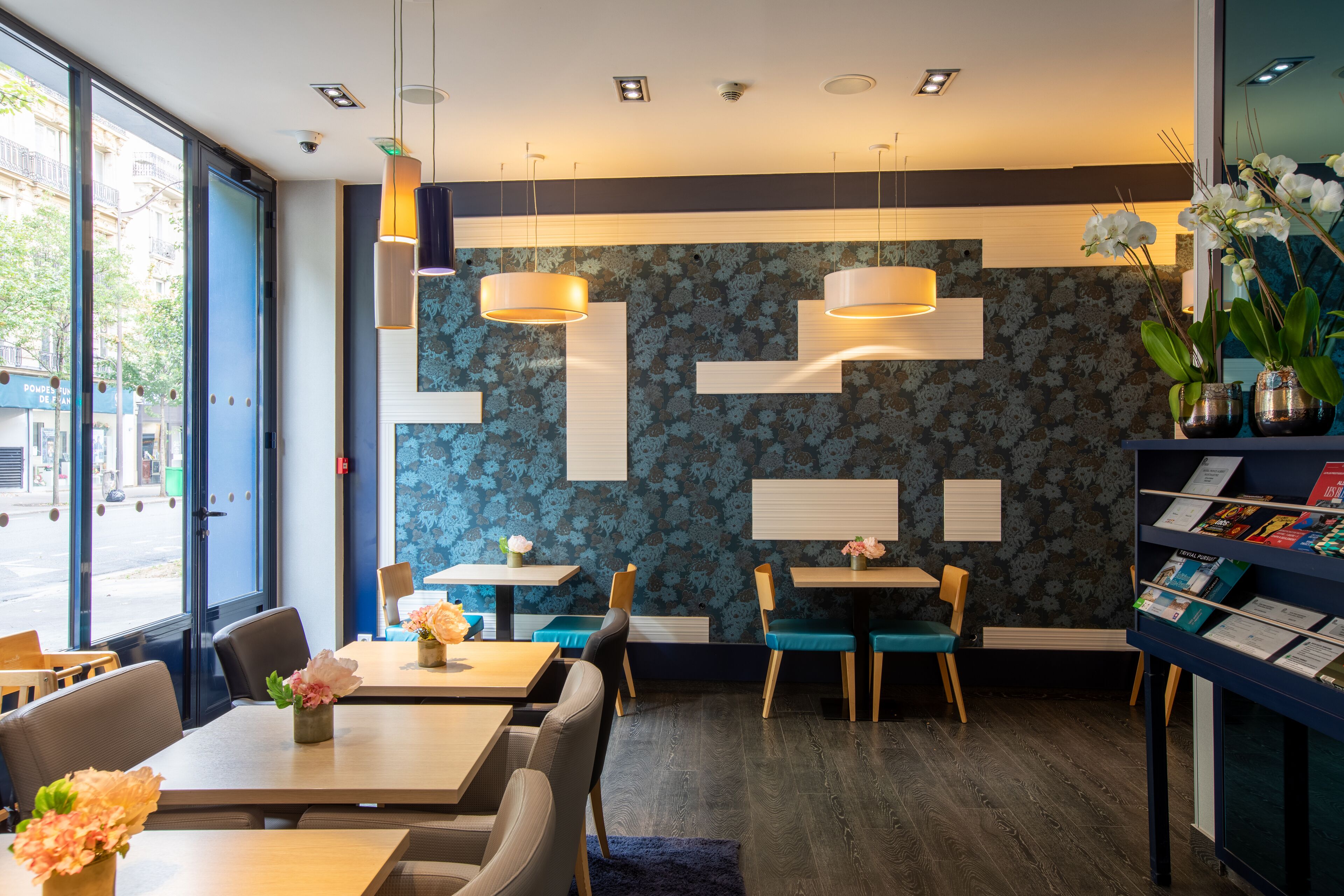 Interior moderno de restaurante con mesas y sillas, iluminación colgante y una estantería con revistas.