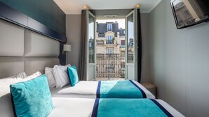 Habitación de hotel con una cama grande, dos almohadas, una mesita de noche y un televisor montado en la pared, además de un balcón con vistas a edificios.
