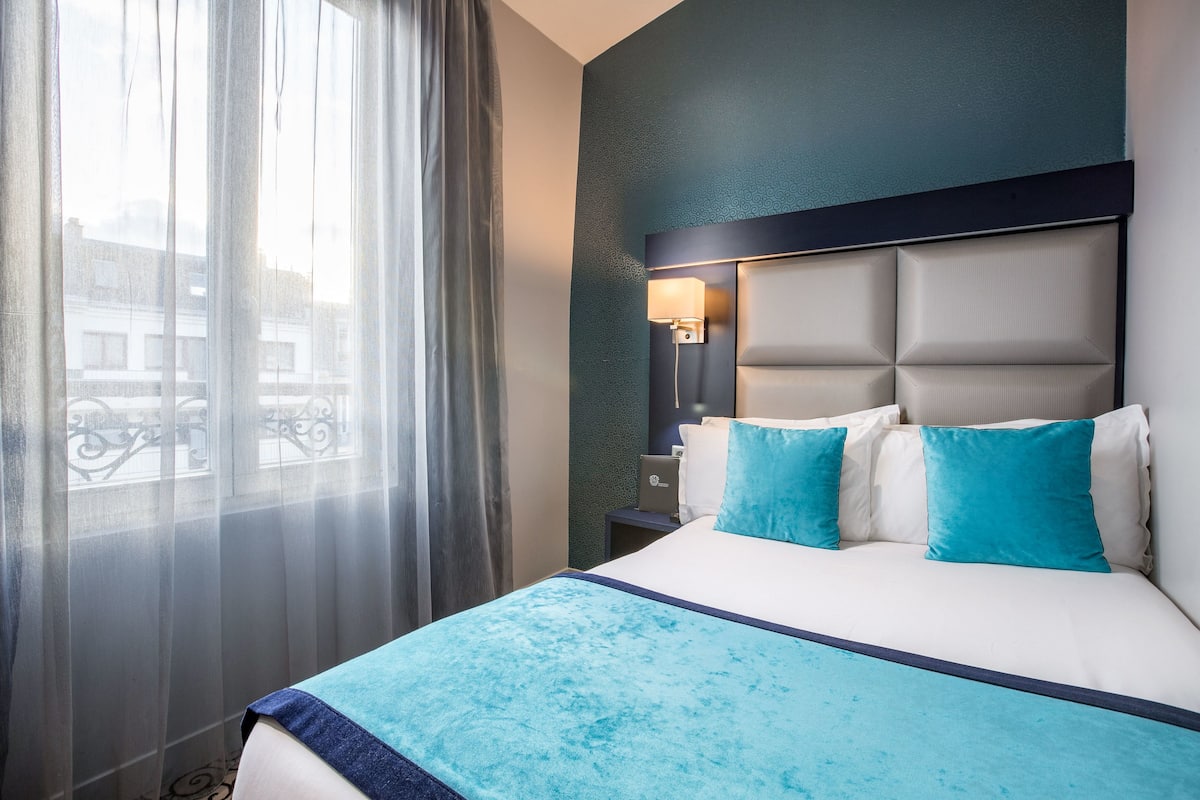 A hotel room with a bed, a nightstand, a lamp, and a view through a window.