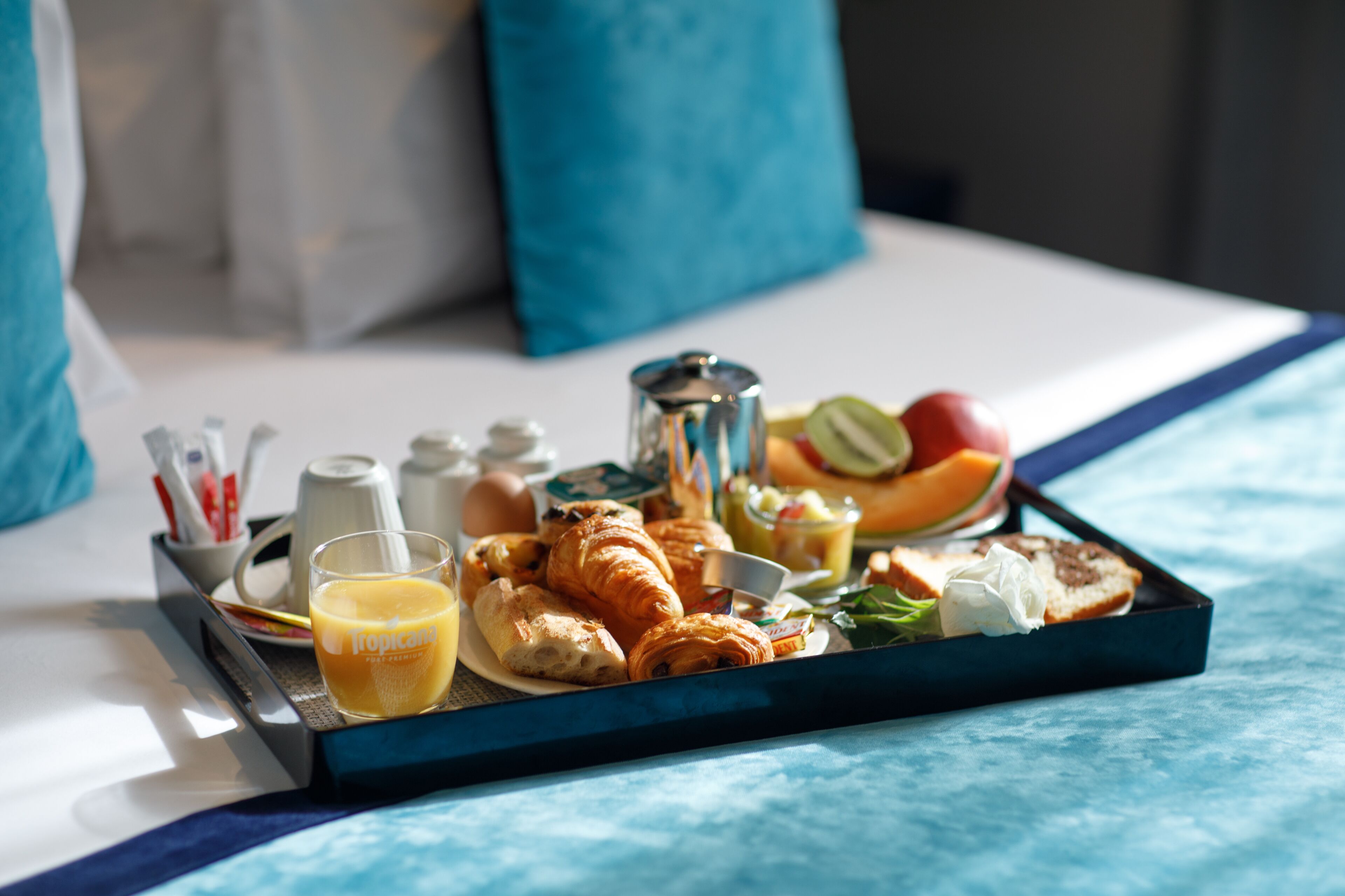 A breakfast tray with croissants, orange juice, and fresh fruit on a bed with blue bedding and pillows.