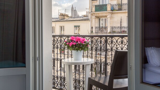 Un balcón con una mesa blanca y una silla, una planta en maceta con flores rosas, y vistas de edificios parisinos.