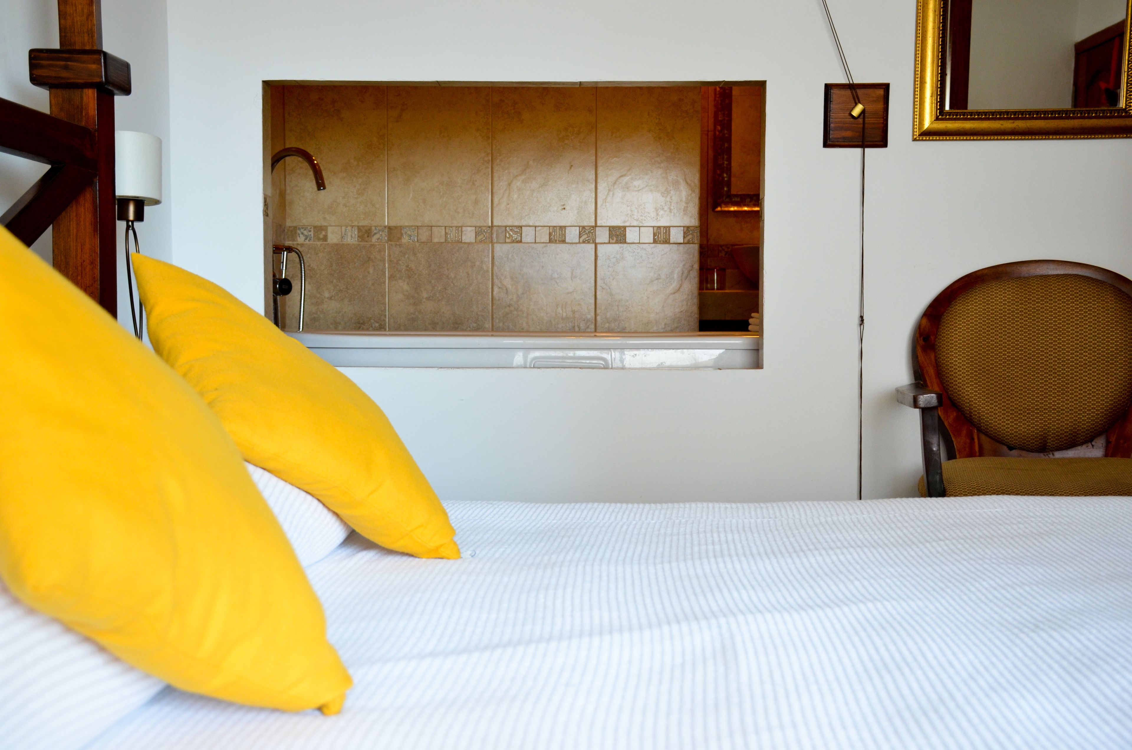 A hotel room with a bed, a window with a view of a bathroom, a chair, and a wall-mounted clock.