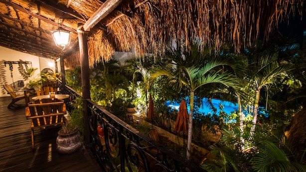 Una terraza de madera con sillas y mesas, rodeada de exuberante vegetación tropical y una estructura de techo de paja.