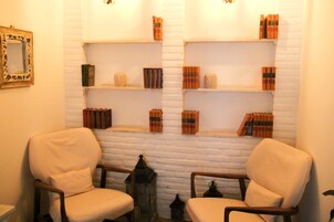 Una habitación con dos sillones, una estantería llena de libros y objetos decorativos, y un espejo en la pared.