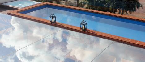Uma piscina retangular com borda revestida de azulejos, duas lanternas decorativas e um reflexo nítido de nuvens na superfície.