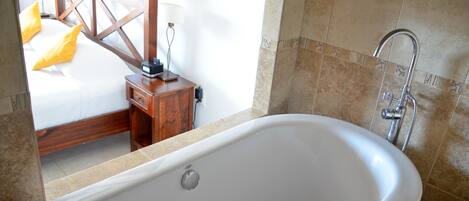 A bathroom with a large bathtub, a wooden nightstand, and a bed with yellow pillows.