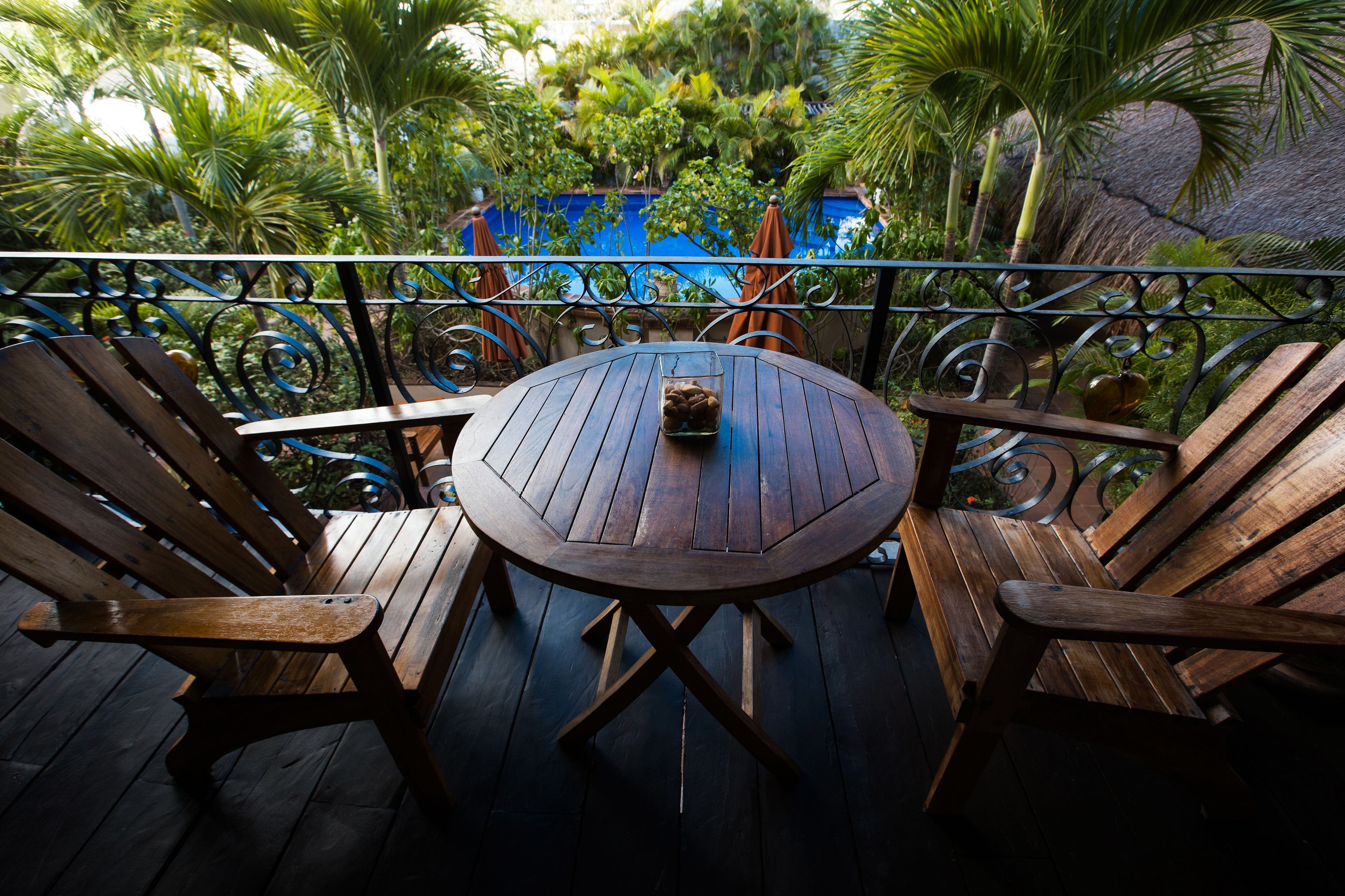Una terraza de madera con una mesa redonda y dos sillas, con vista a una piscina y abundante vegetación.