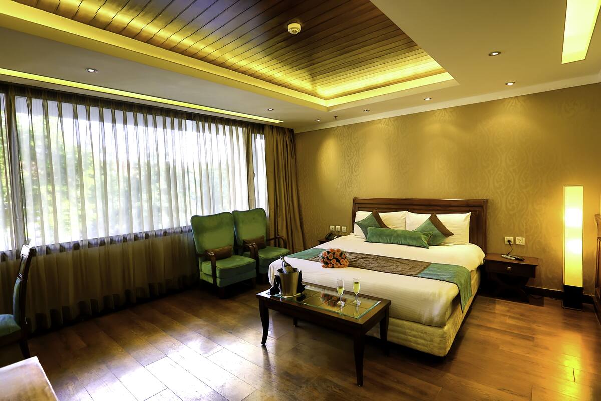 A hotel room with a large bed, green chairs, a wooden coffee table, and a nightstand with a lamp.