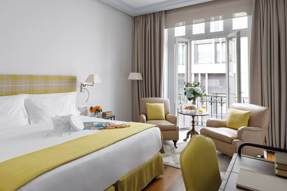 Quarto de hotel com uma cama grande, duas poltronas, uma mesinha com um vaso de flores e vista da cidade através de grandes janelas.