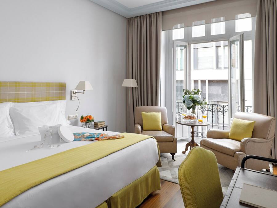 Habitación de hotel con una cama grande, dos sillones, una mesita con un florero y vistas a la ciudad a través de amplios ventanales.