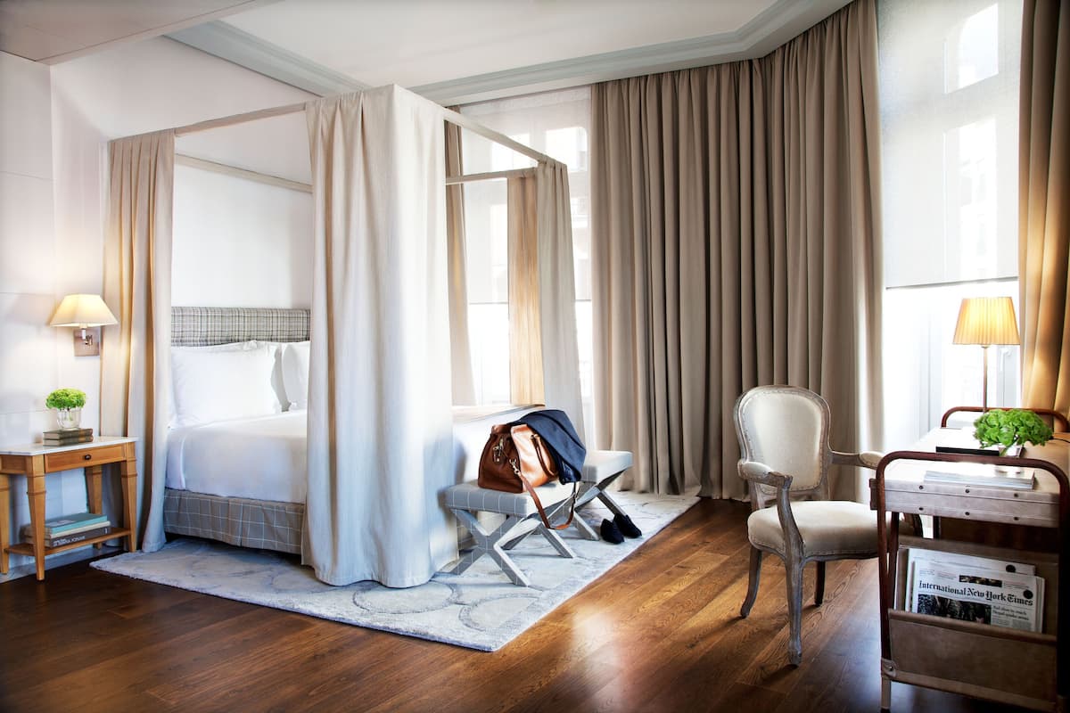Quarto de hotel com cama de dossel, uma mesa com cadeira, uma mesinha de canto com abajur e uma janela com cortinas.