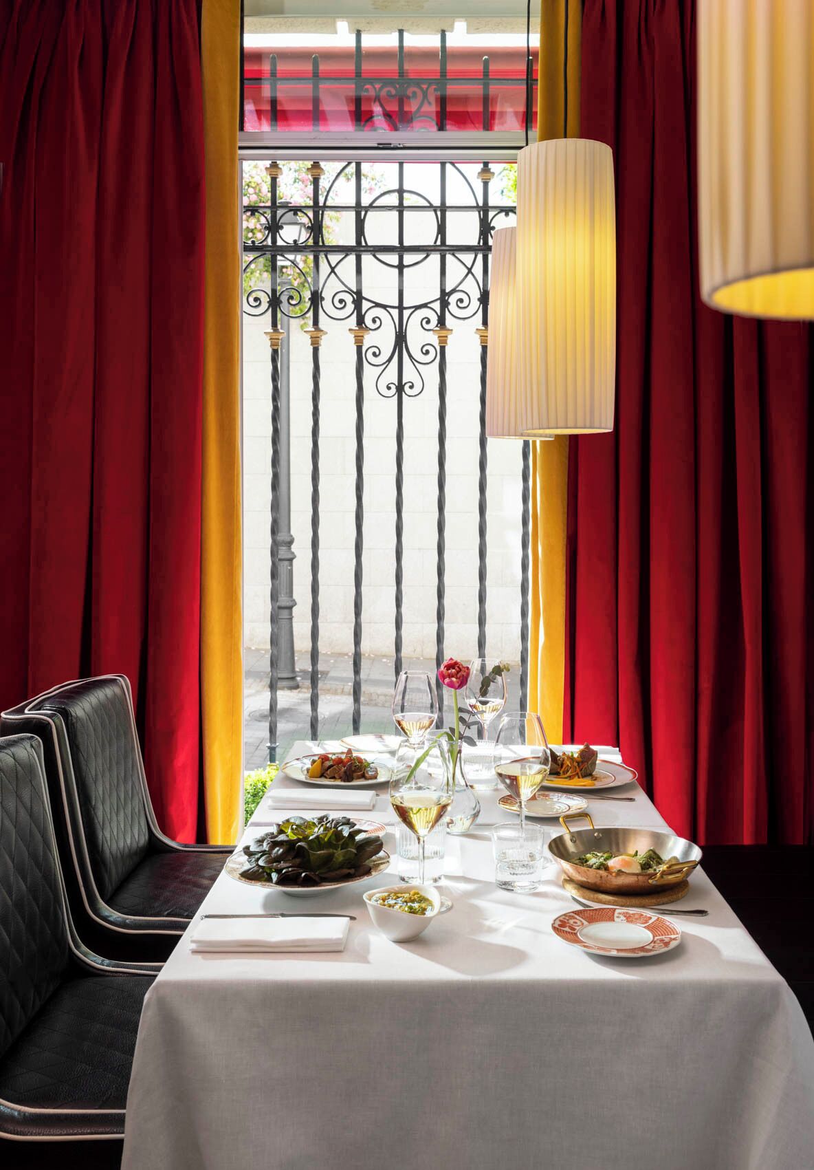 Une table à manger dressée avec des assiettes, des verres et un vase contenant une fleur, avec des rideaux rouges et une vue sur un balcon en arrière-plan.