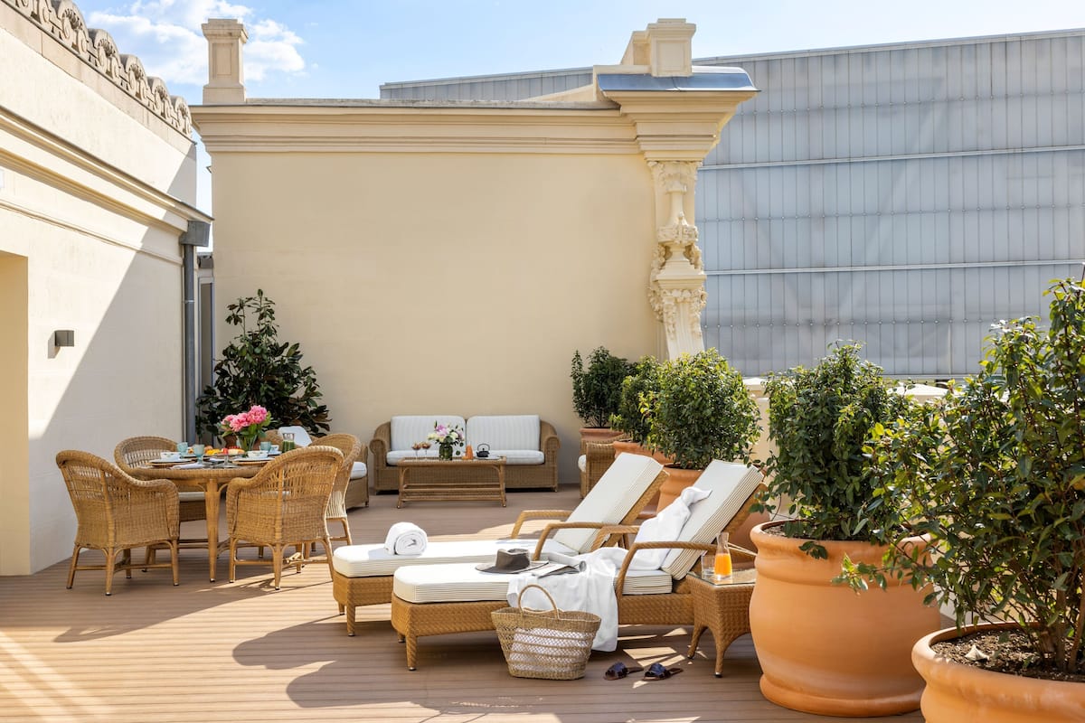Área de estar ao ar livre com boa iluminação, móveis de vime, plantas em vasos e vista para um edifício ao fundo.