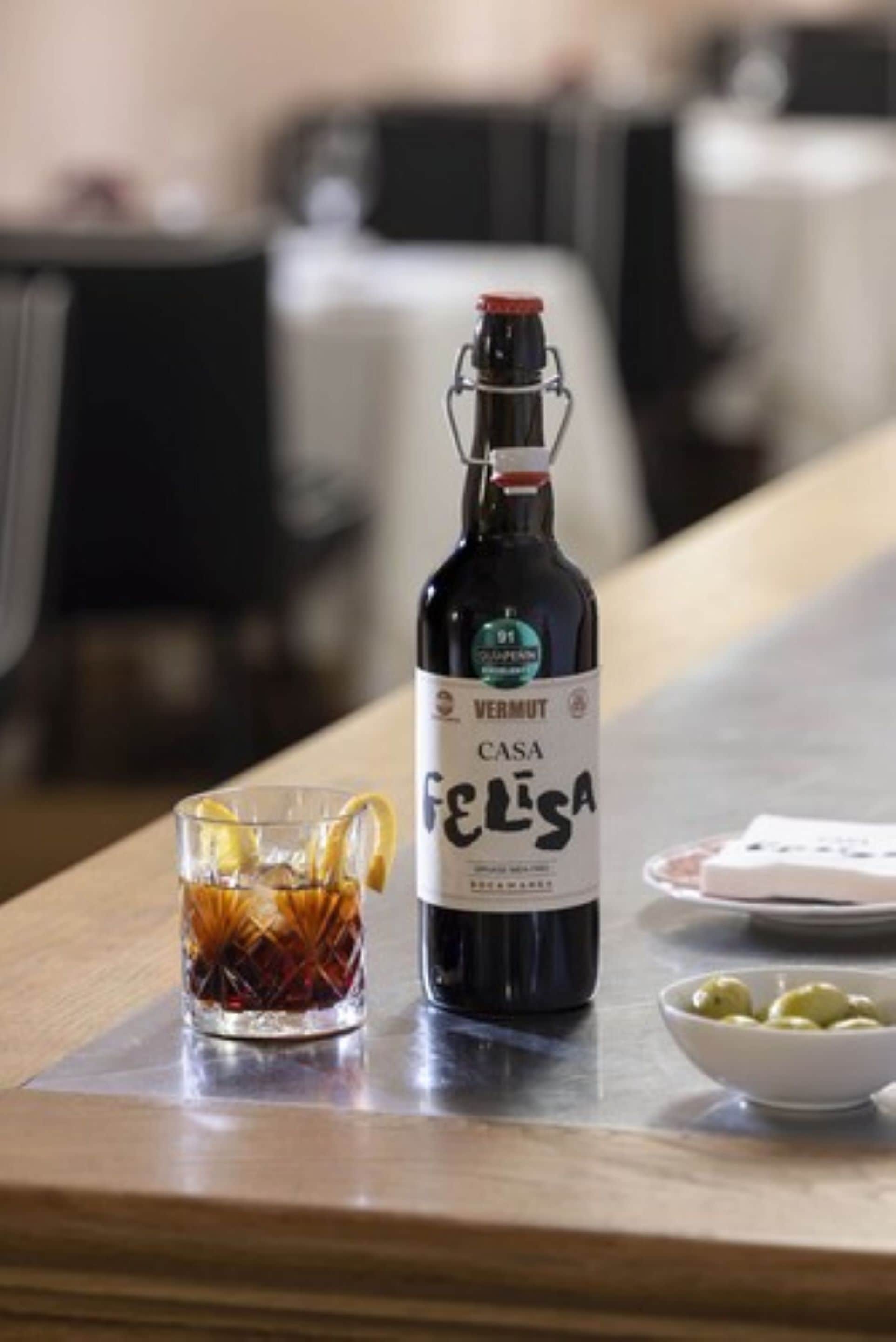A bottle of Vermut Casa Felisa wine, a glass with a lemon slice, and a bowl of olives on a wooden table in a dining area.