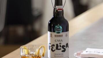 A bottle of Vermut Casa Felisa wine, a glass with a lemon slice, and a bowl of olives on a wooden table in a dining area.