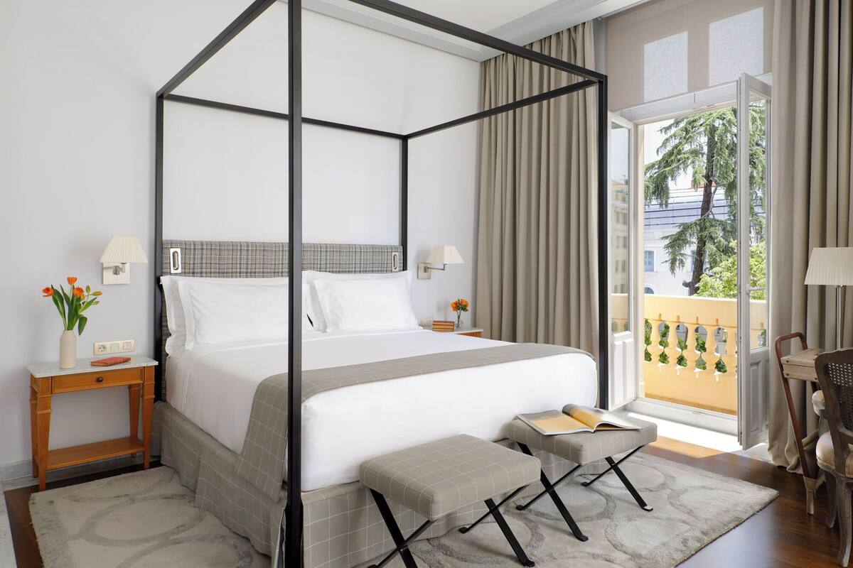 Uma cama de dossel com cabeceira xadrez, uma mesinha de cabeceira com abajur, um pequeno banco e vista para o exterior através de uma janela grande.