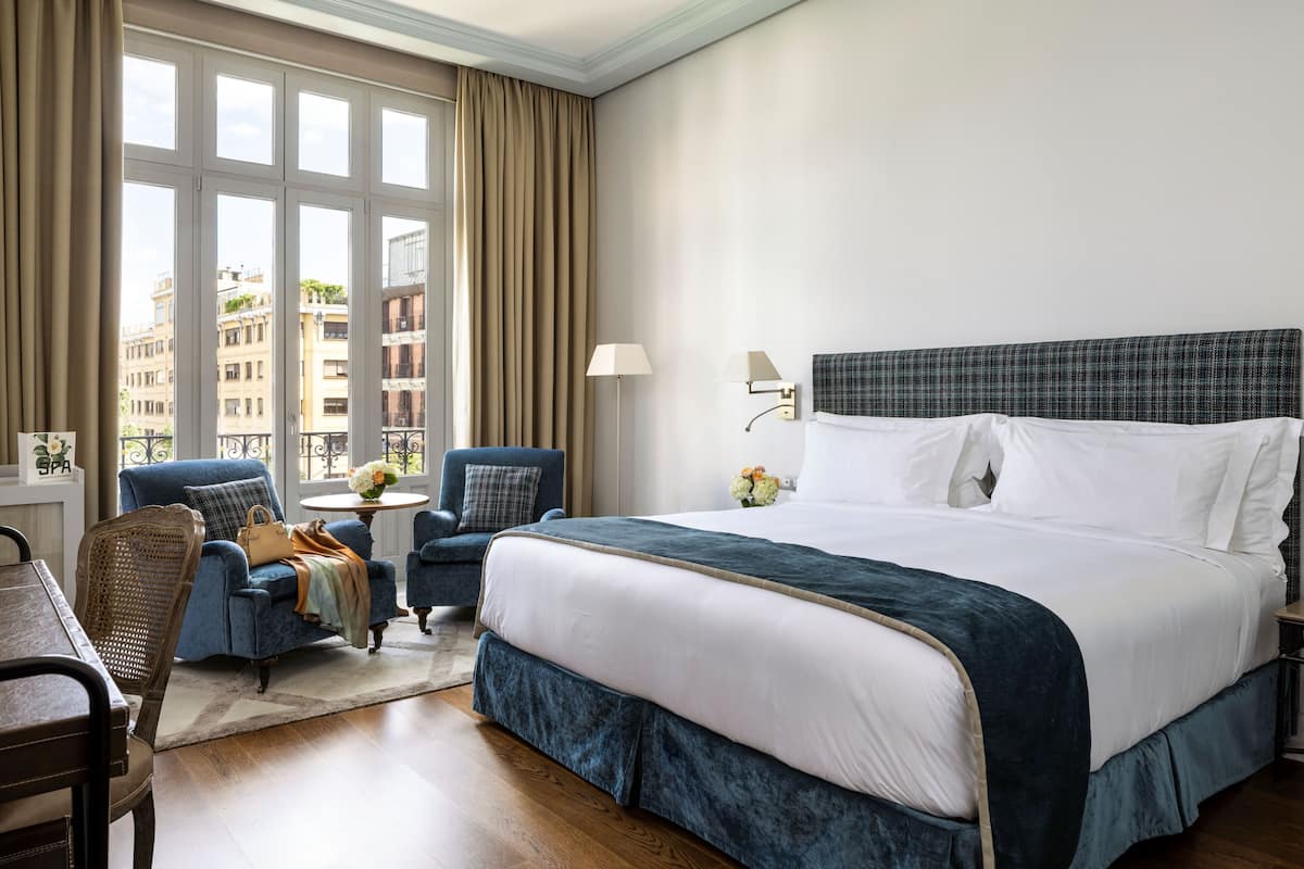 Quarto de hotel com uma cama grande, duas poltronas, uma mesa pequena com um vaso de flores e vista para edifícios através de grandes janelas.