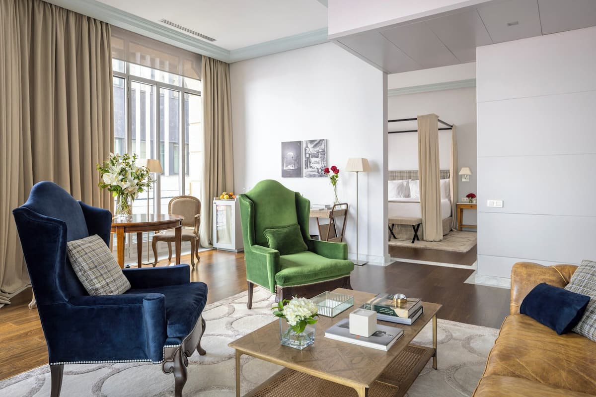 Uma sala de estar espaçosa com uma poltrona azul, uma poltrona verde e um sofá marrom. Há uma mesa de centro com um vaso de flores e livros. Uma área de jantar com mesa e cadeiras é visível ao fundo.