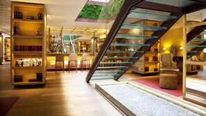 Un interior moderno con una escalera de cristal, estanterías de madera con libros y una zona de estar con un reposapiés estampado.