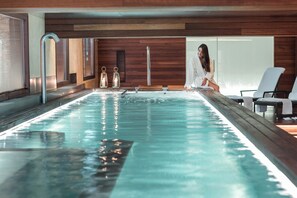 A modern indoor swimming pool with wooden paneling, a seating area with white chairs, and a person in a robe. - URSO Hotel & Spa (Madrid)
