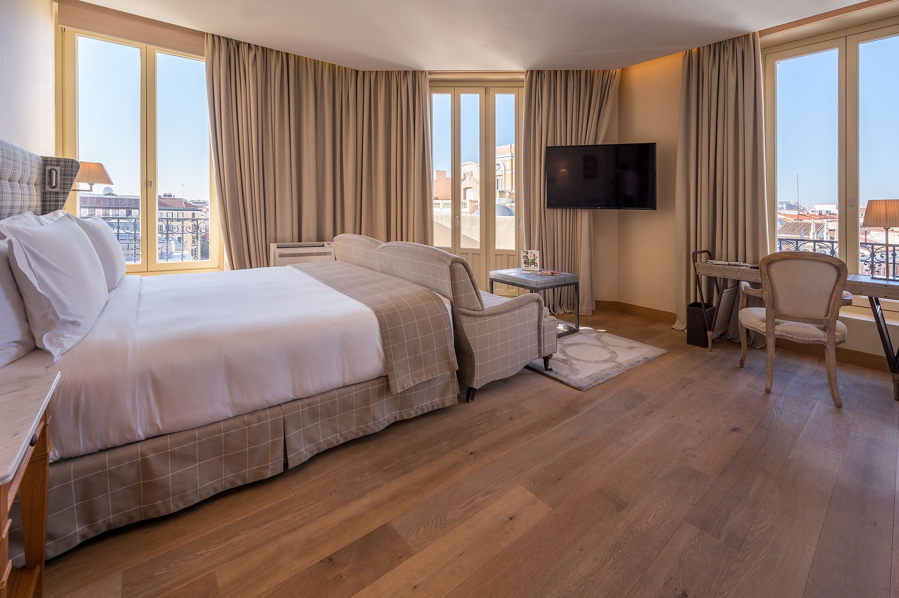 A hotel room with a large bed, a desk, a chair, a TV, and a view of the city through large windows.