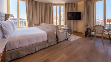 A hotel room with a large bed, a desk, a chair, a TV, and a view of the city through large windows.