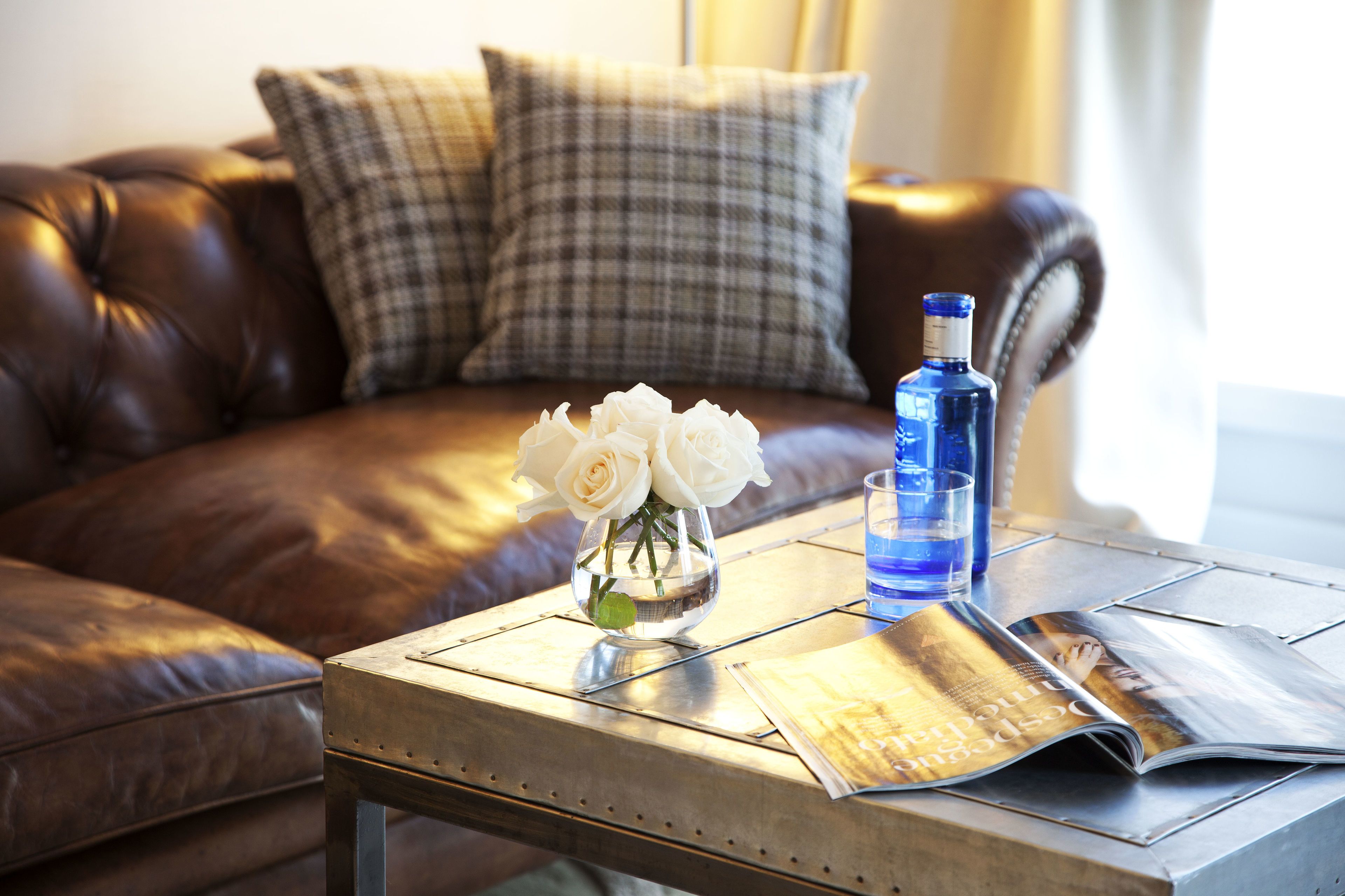 A brown leather sofa with plaid cushions, a glass vase with white roses, a blue bottle, and a magazine on a metal table.
