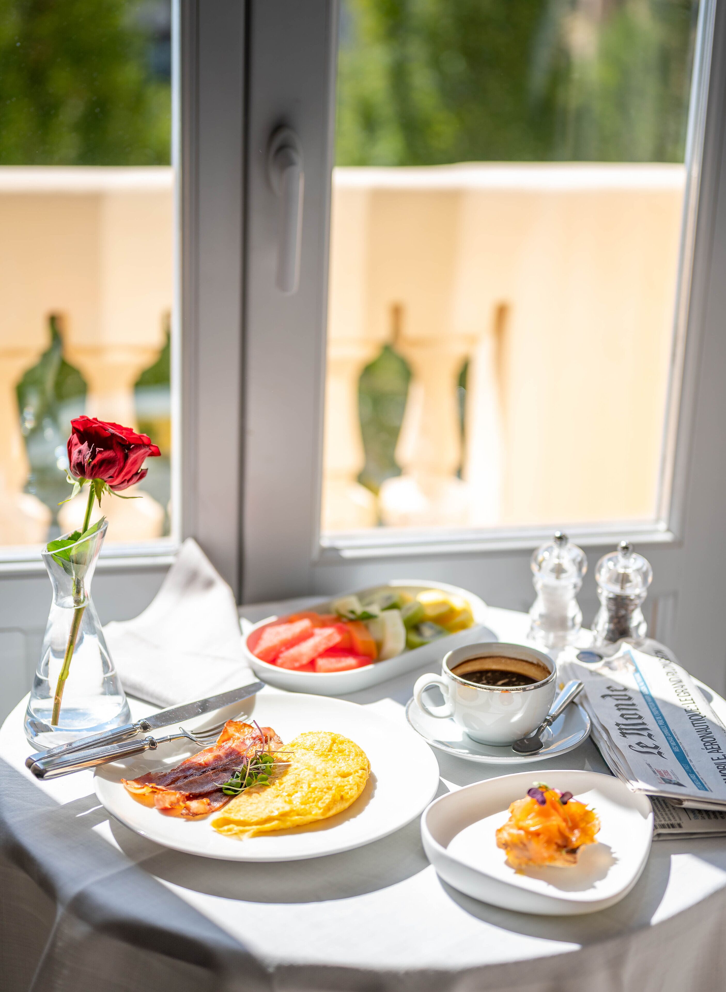 A breakfast table with plates of food, a cup of coffee, a vase with a single rose, and salt and pepper shakers.