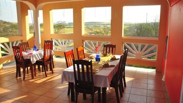 A dining area with a table set for four, surrounded by chairs, large windows with white frames, and a view of a landscape.