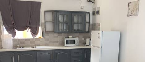 A modern kitchen with grey cabinets, a white refrigerator, a microwave, and a sink. There is a grey sofa in the living area.