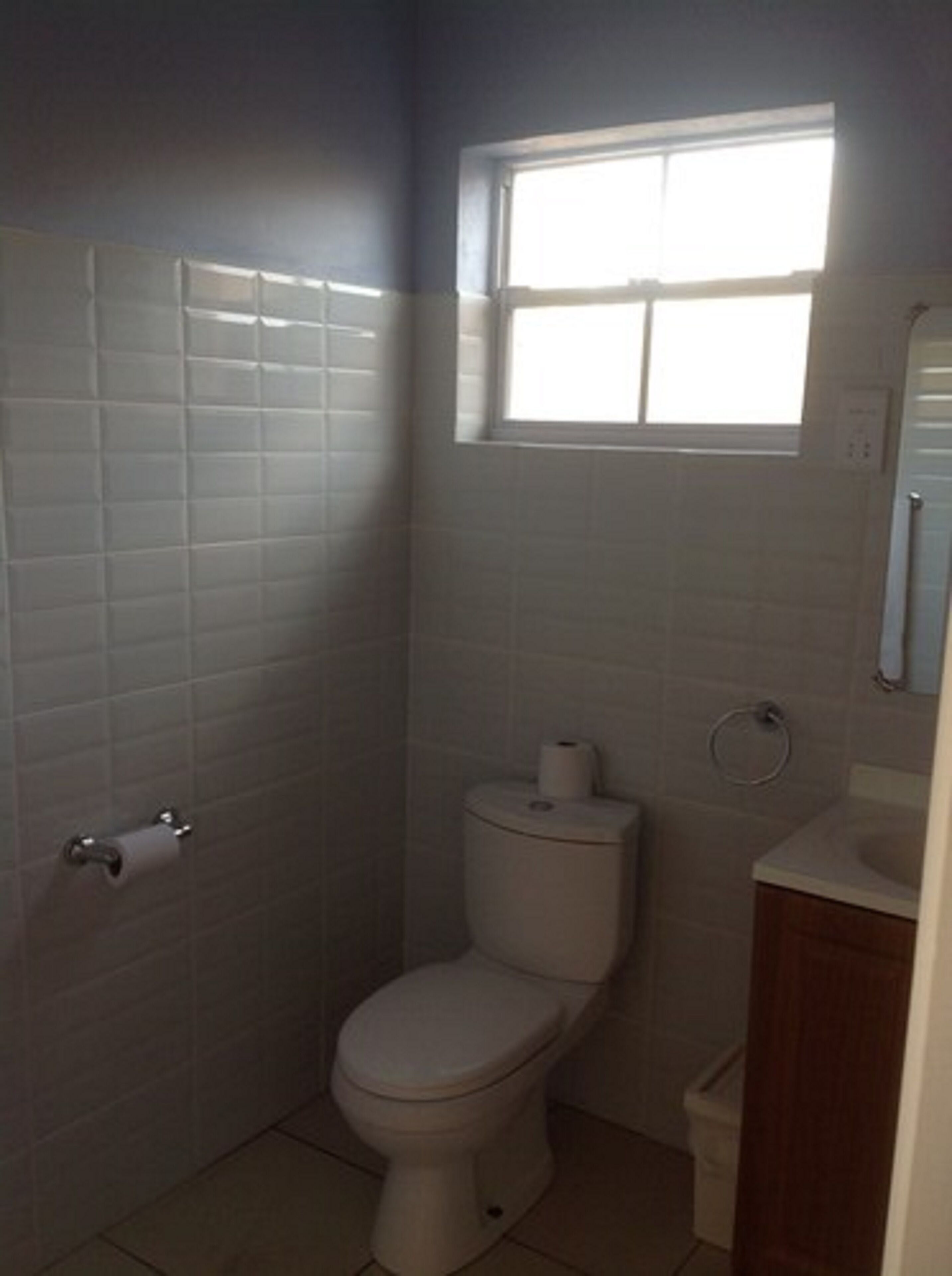 A small bathroom with white tiles, a toilet, a window, and a sink.