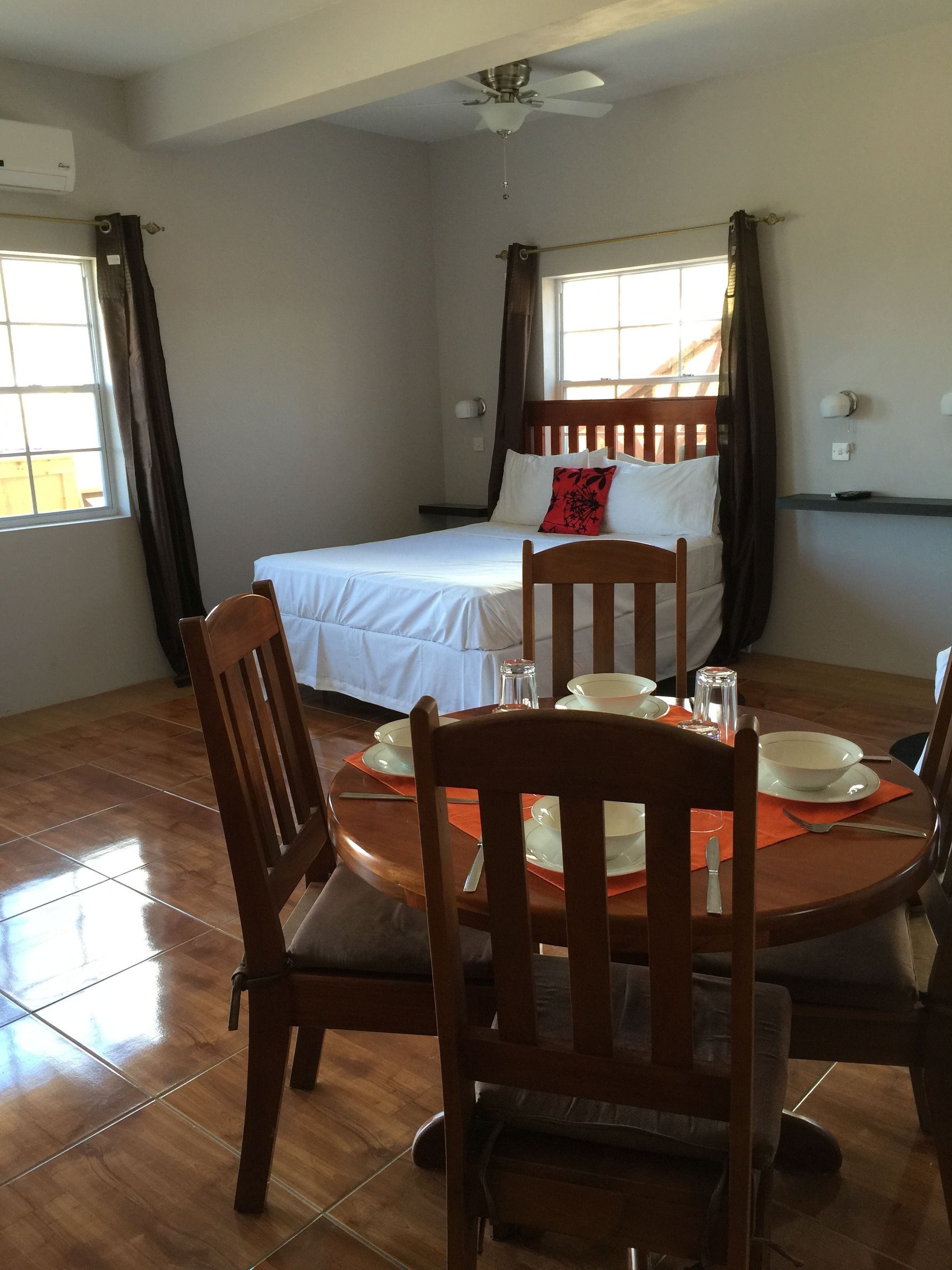 Una habitación con cama, una mesa de comedor con sillas, un ventilador de techo y una ventana con cortinas.