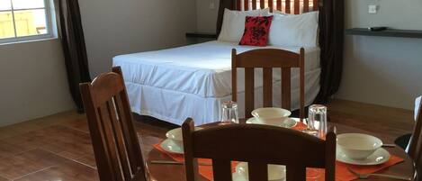 Una habitación con cama, una mesa de comedor con sillas, un ventilador de techo y una ventana con cortinas.
