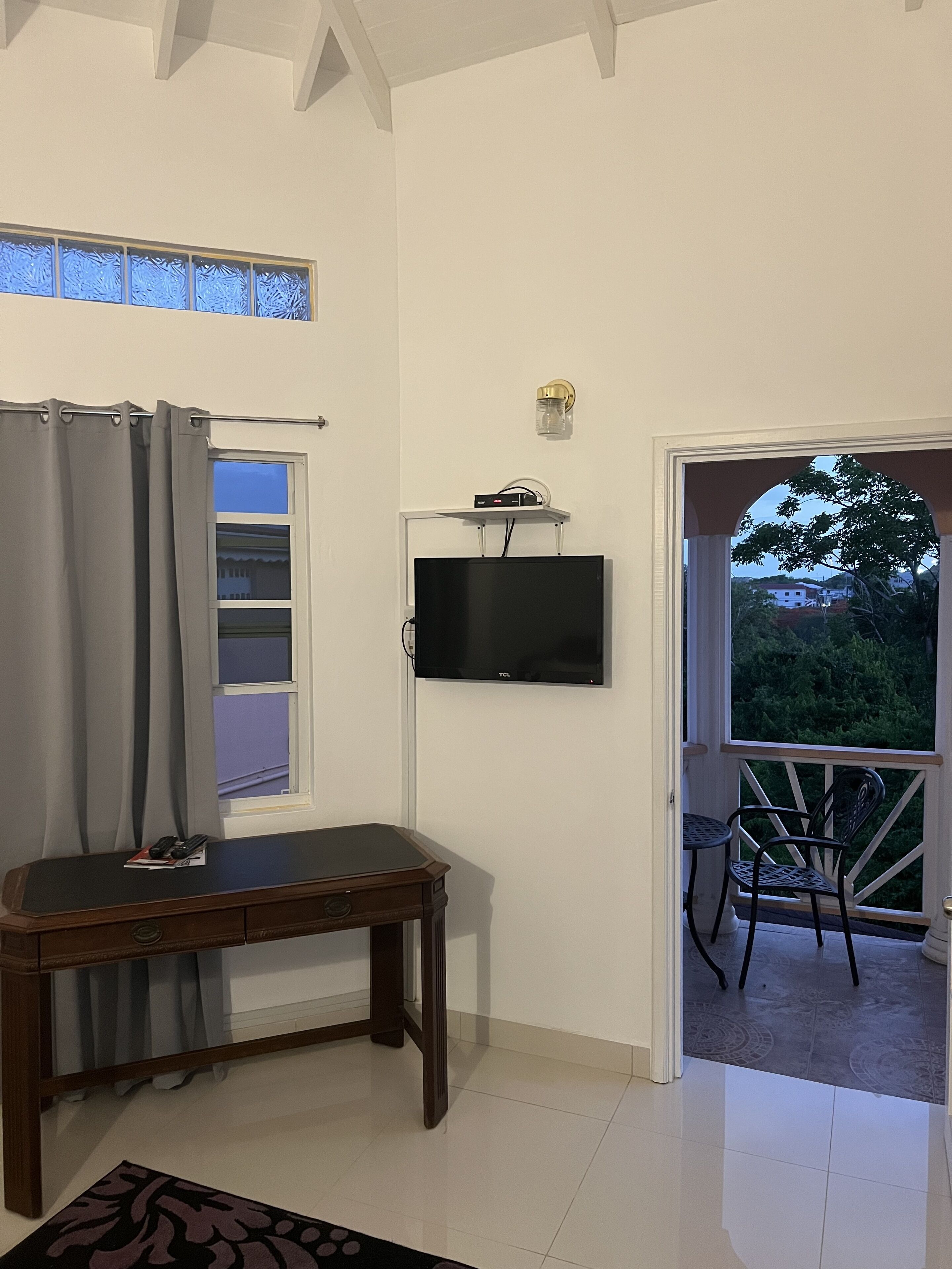 Una stanza con una TV a parete, una scrivania in legno, una finestra con tende e un balcone con sedie.
