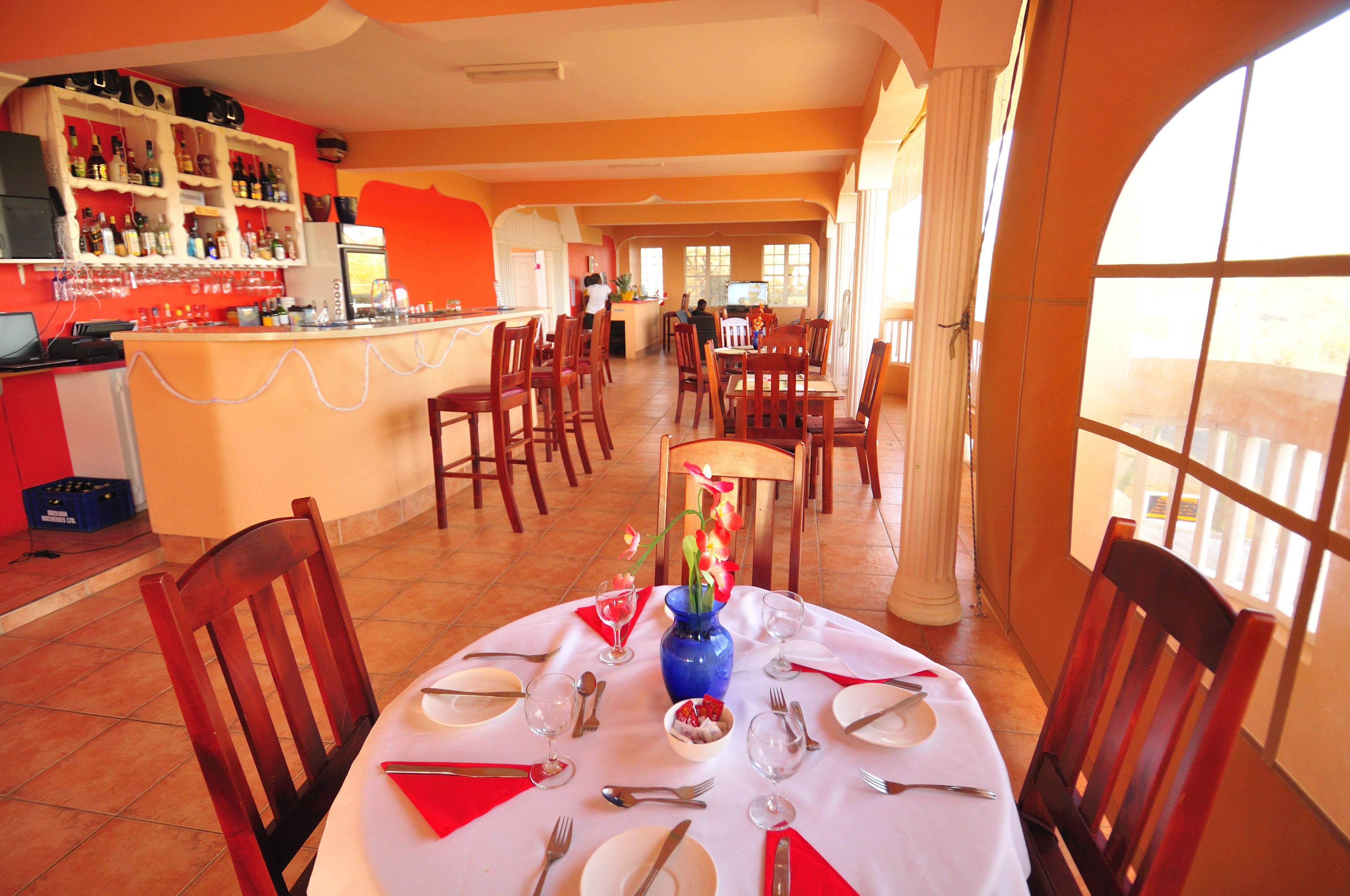 A restaurant with a bar area, multiple dining tables set with red napkins, and a vase with flowers on one table.