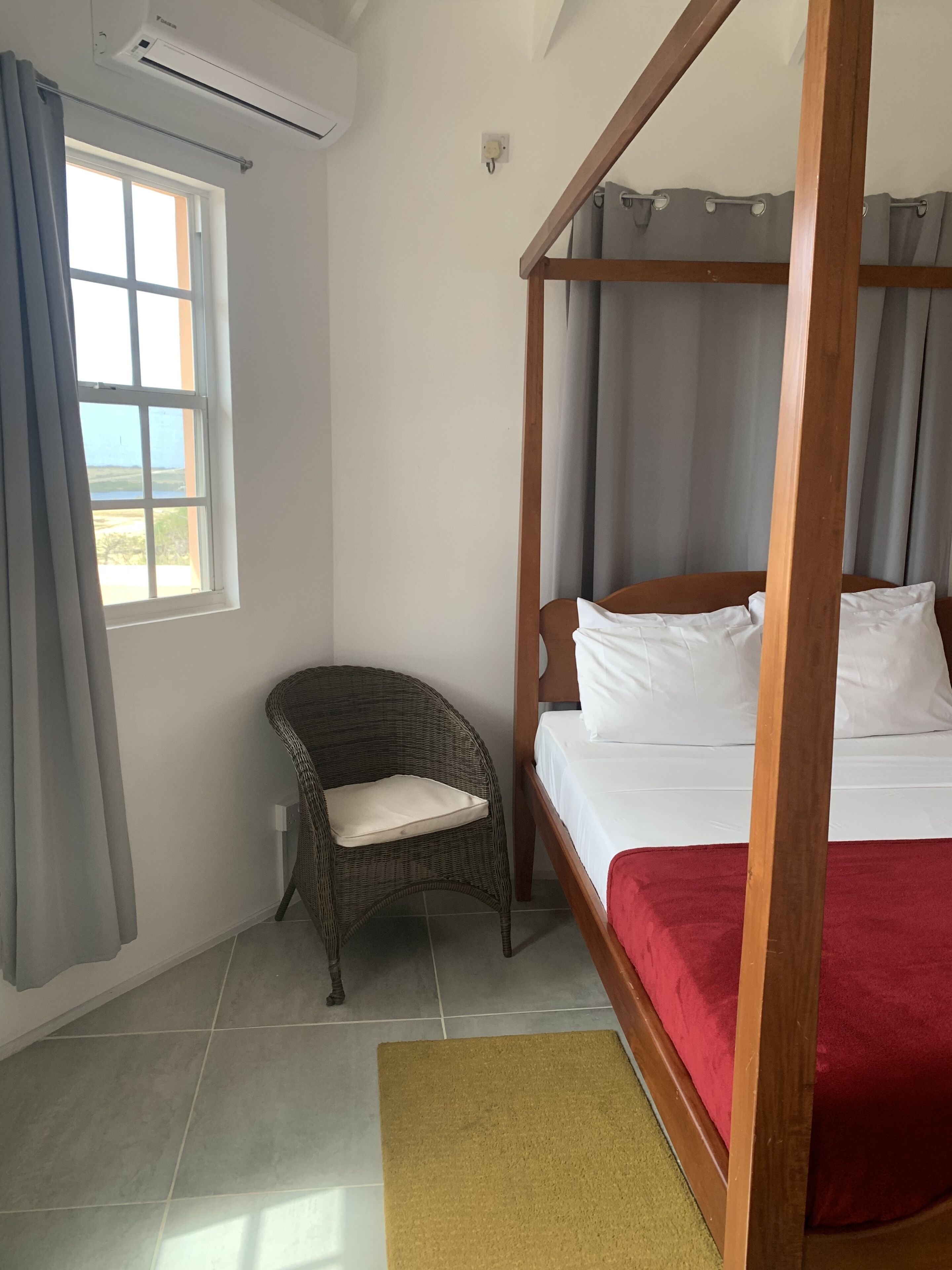 A room with a four-poster bed, a wicker chair, a window with curtains, and a tiled floor.