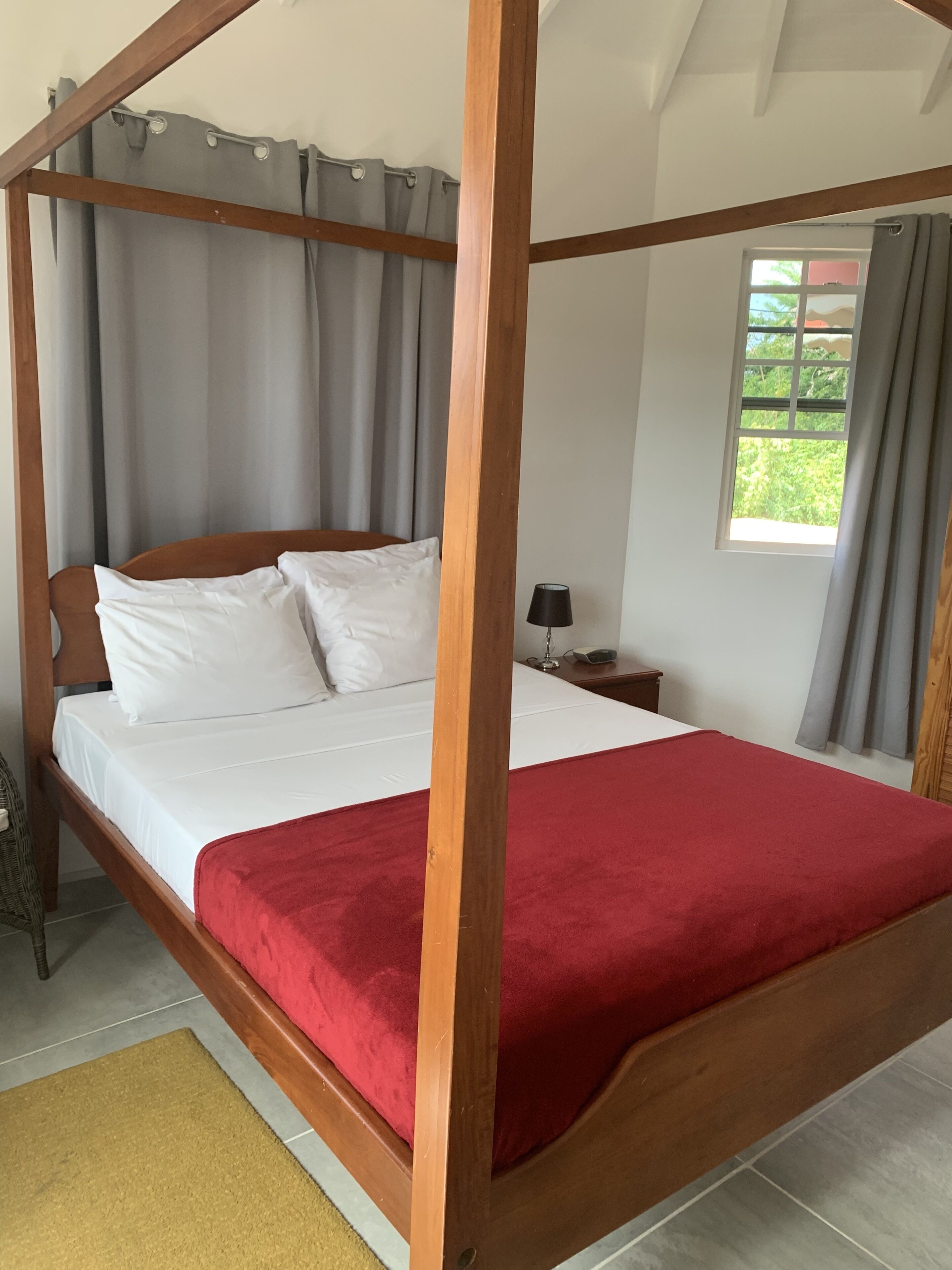 A four-poster bed with white linens and a red throw blanket, a bedside table with a lamp, and a window with curtains.