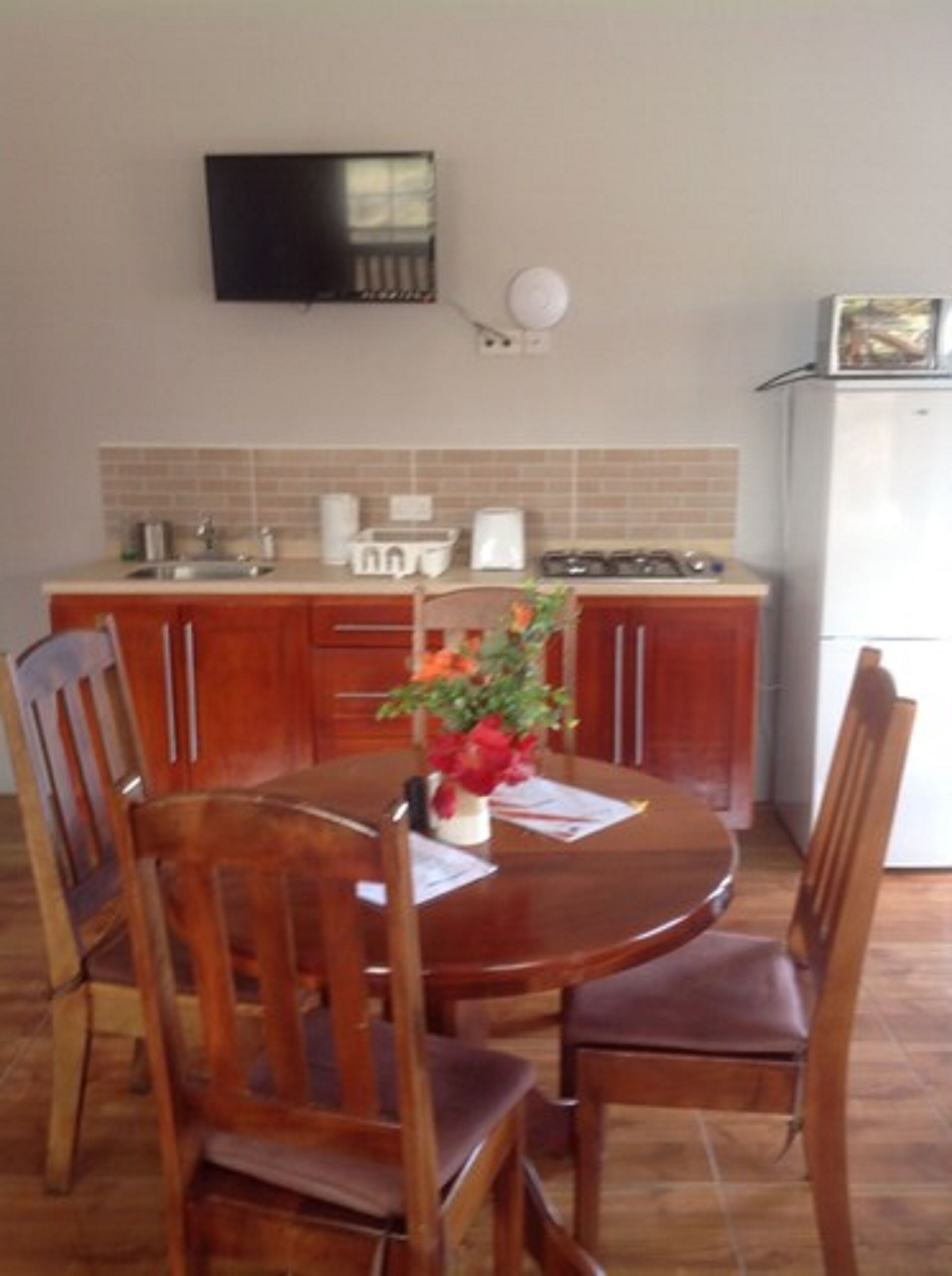 Una cocina con una mesa de comedor, sillas, un televisor montado en la pared y un refrigerador.