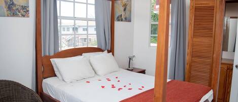 A four-poster bed with white linens and a red throw blanket, a wicker chair, and a nightstand with a lamp.