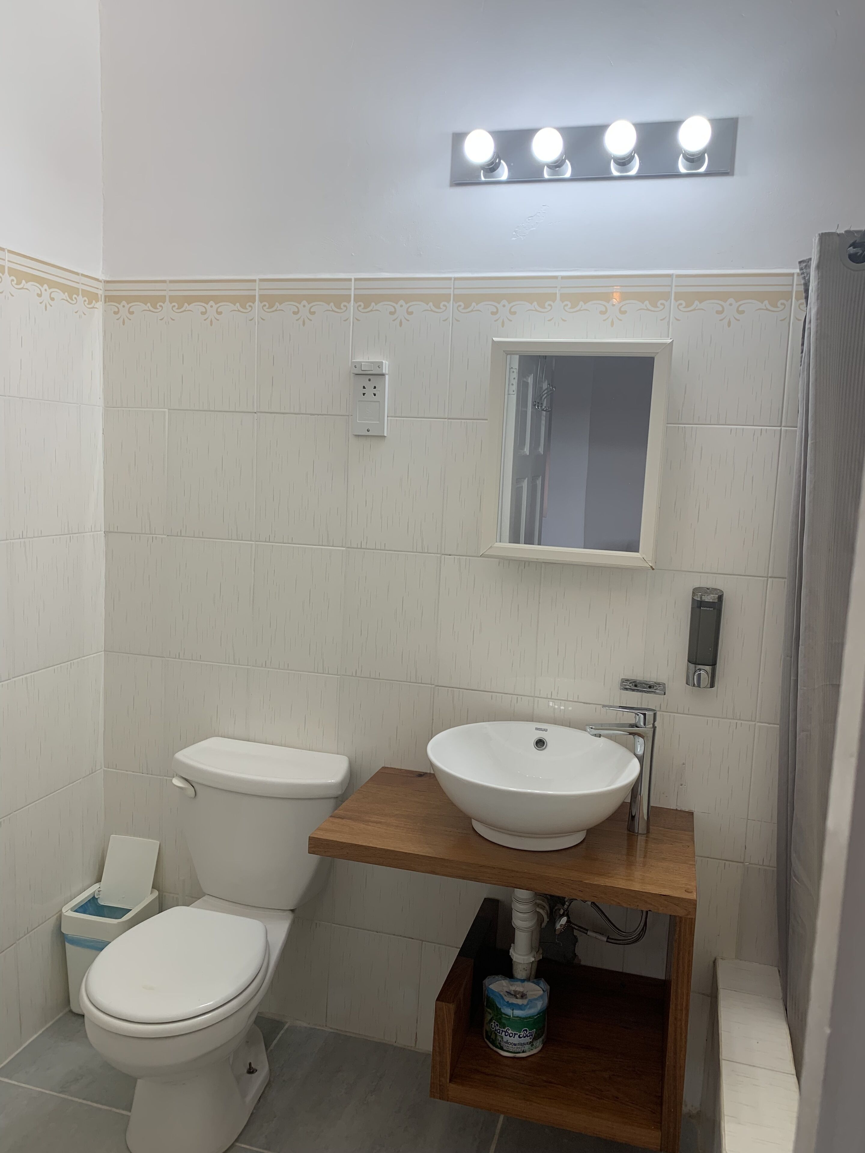 Ein Badezimmer mit Toilette, Waschbecken und Spiegel. Über dem Spiegel befindet sich eine Leuchte und an der Wand ein Seifenspender.