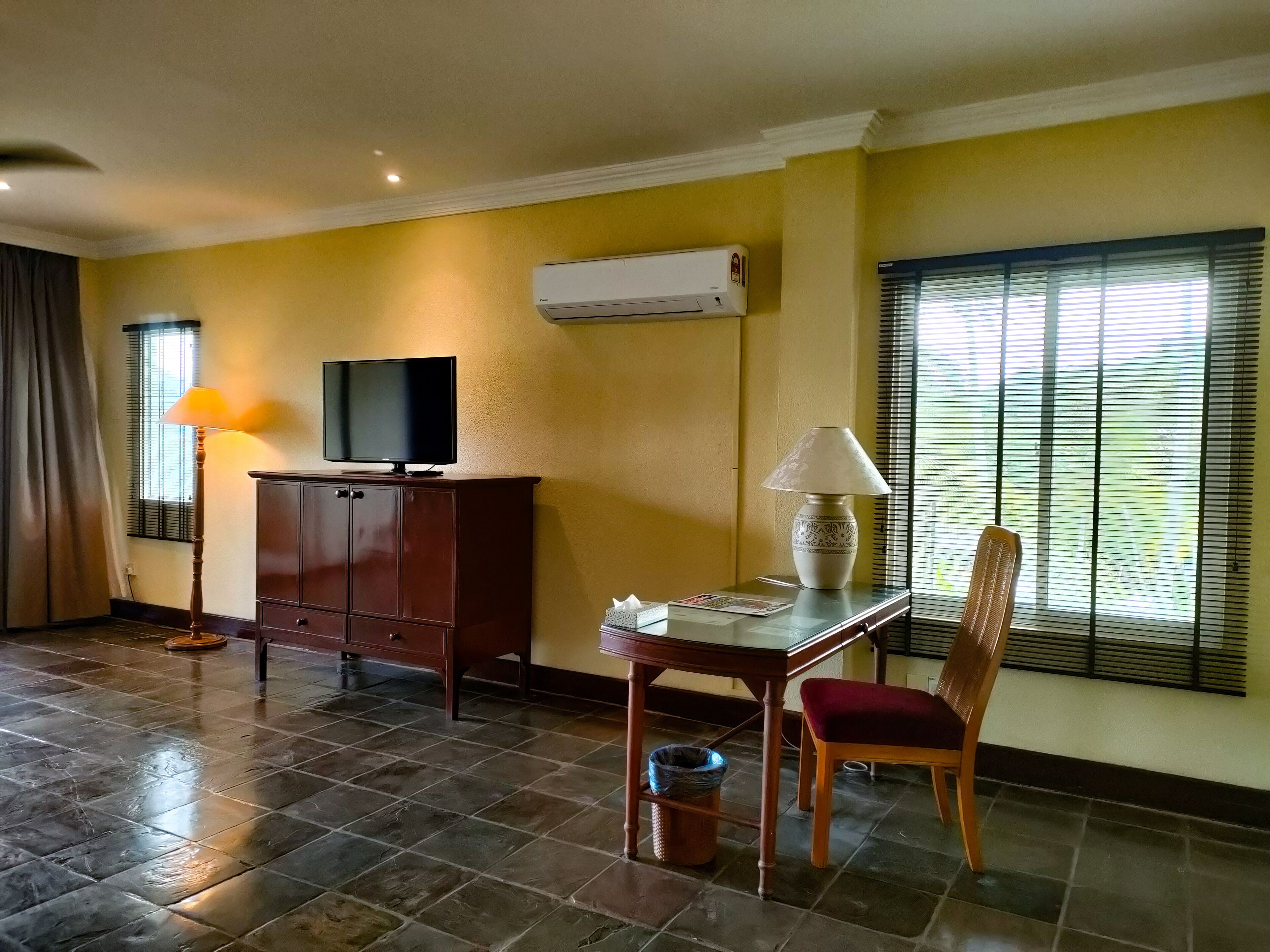 A hotel room with a TV on a cabinet, a table with a lamp, a chair, and a window with blinds.