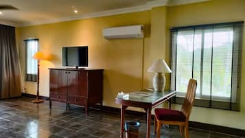 A hotel room with a TV on a cabinet, a table with a lamp, a chair, and a window with blinds.