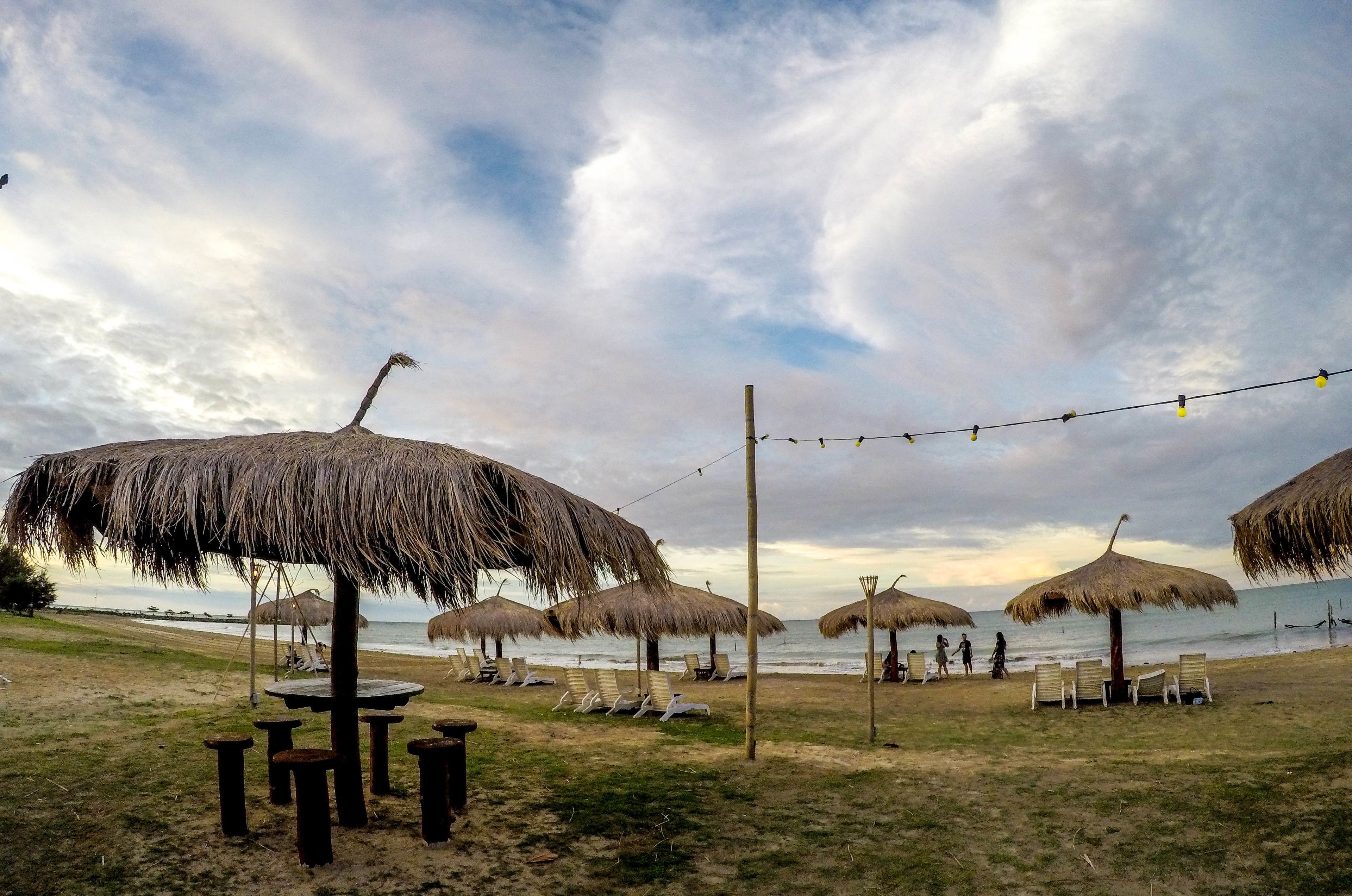 A beach with thatched umbrellas and lounge chairs, a string of lights, and a view of the sea.