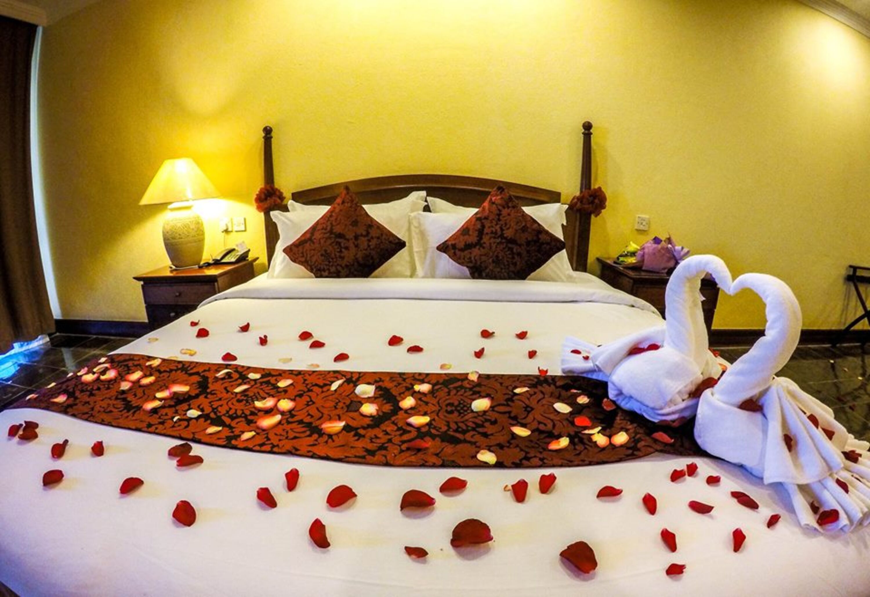 Une chambre d’hôtel avec un lit décoré de pétales de roses et de serviettes en forme de cygne, une lampe de chevet et une couvre-lit à motifs floraux.