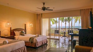 A hotel room with two beds, a ceiling fan, a small table, and a view of the ocean through large windows.