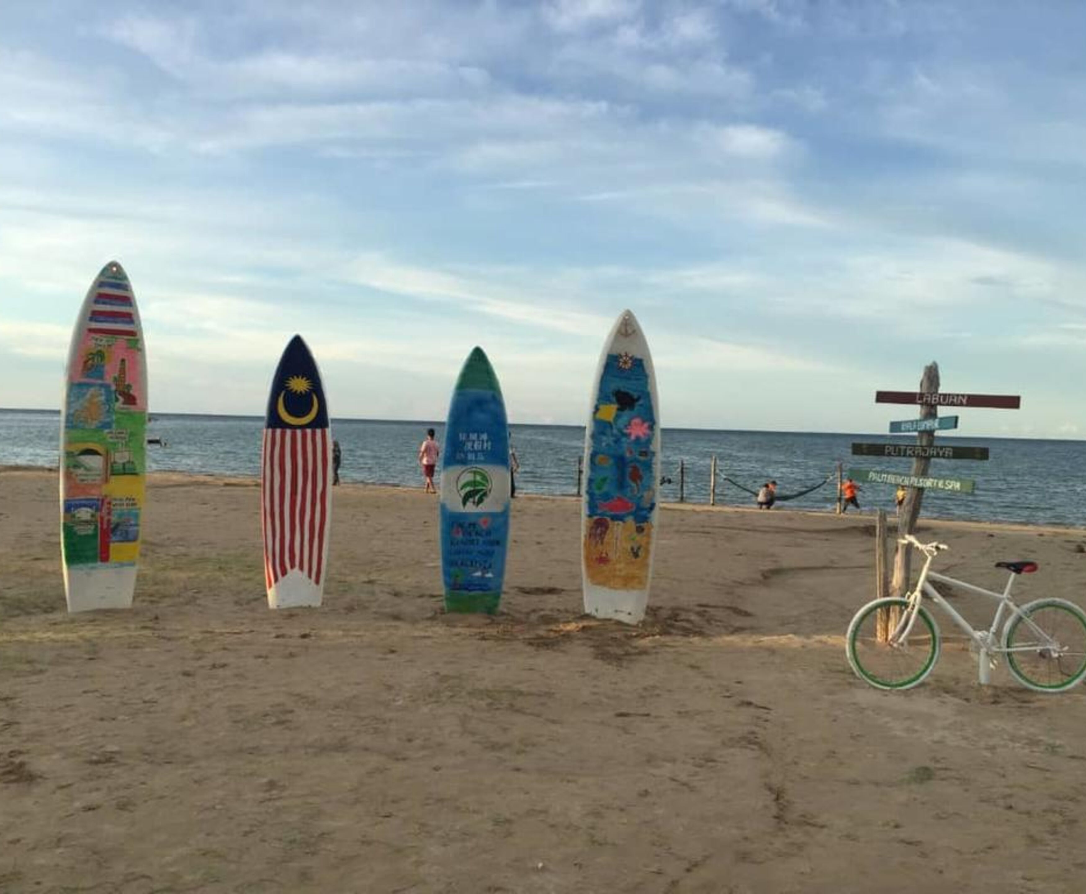 Surfboards with various designs and a bicycle on a sandy beach with a signpost in the background.
